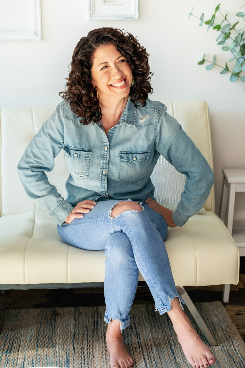 Smiling woman sitting on a cream colored with her hands on her hips and legs crossed in a bright room.