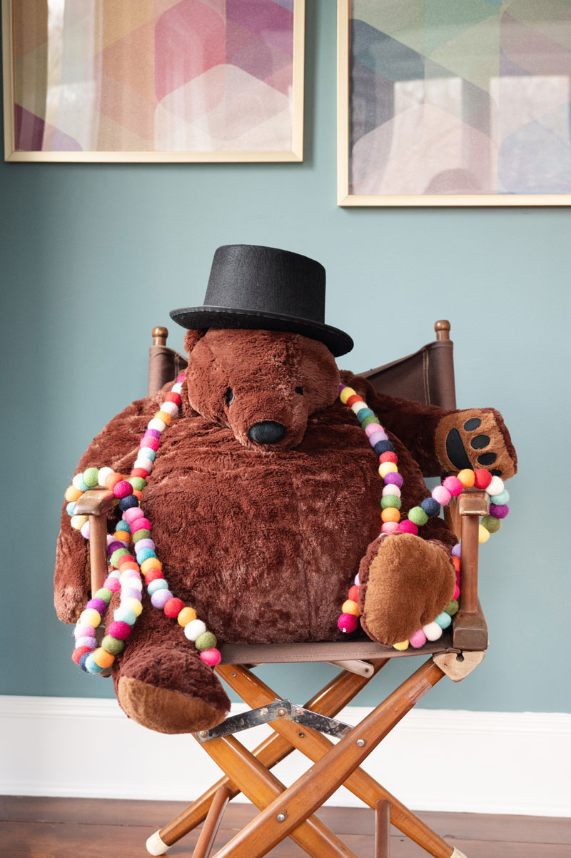 A giant brown teddy bear is sitting in a directors chair wearing a black top hat and strand of felt ball garland around its neck.
